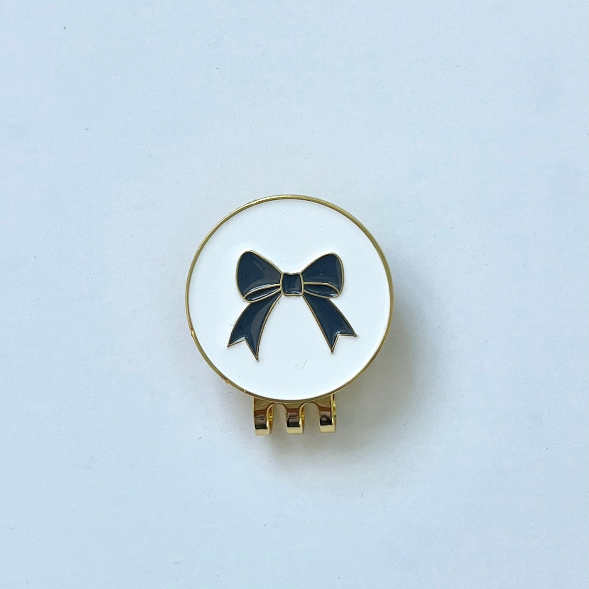 A gold-tone metal golf ball marker with a white enamel-filled bow on a white background, designed to be attached to a hat.