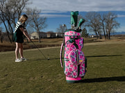 Woman practicing golf on a course with a pink floral golf bag.
